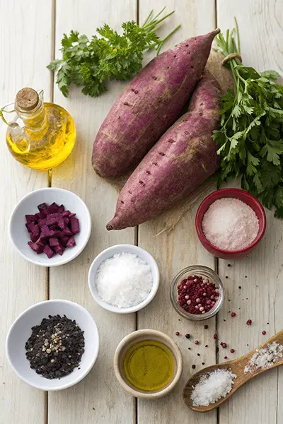 Ingredients for a purple sweet potato recipe, including whole purple sweet potatoes, herbs, spices, and olive oil arranged on a wooden table.