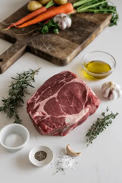 Ingredients for a bottom round roast recipe, including raw beef roast, fresh herbs, garlic, olive oil, salt, pepper, and vegetables on a wooden board.