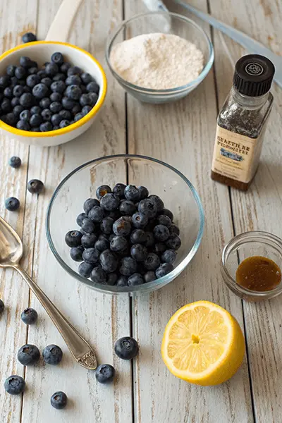 A collection of ingredients for making blueberry compote recipe, including fresh blueberries in a glass bowl, granulated sugar, a halved lemon with zest, and vanilla extract, arranged on a rustic wooden table.