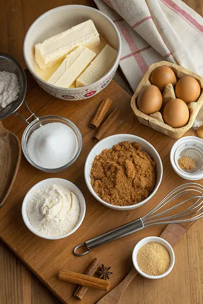 A wooden board with ingredients for churro cheesecake recipe, including cream cheese, eggs, brown sugar, white sugar, flour, cinnamon sticks, and butter.