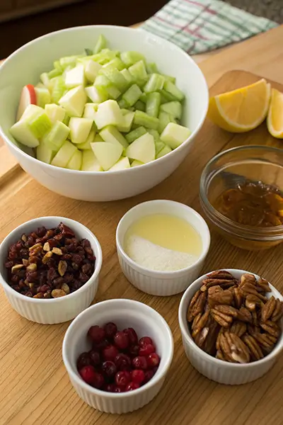 Ingredients for apple salad including chopped apples, celery, lemon juice, pecans, cranberries, raisins, and dressing in small bowls.