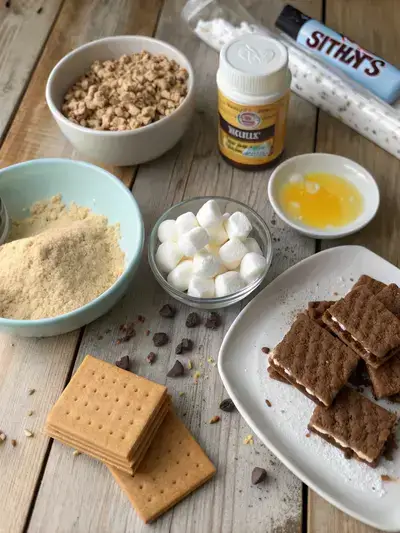 Ingredients for S'mores Bars displayed on a kitchen table, including graham cracker crumbs, sugar, butter, marshmallows, chocolate chips, and cookies.